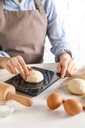 Photo of Woman weighing raw dough ball on kitchen scale at white wooden table, closeup