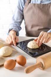 Photo of Woman weighing raw dough ball on kitchen scale at white wooden table, closeup