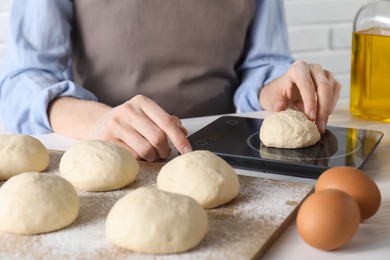 Photo of Woman weighing raw dough ball on kitchen scale at white wooden table, closeup