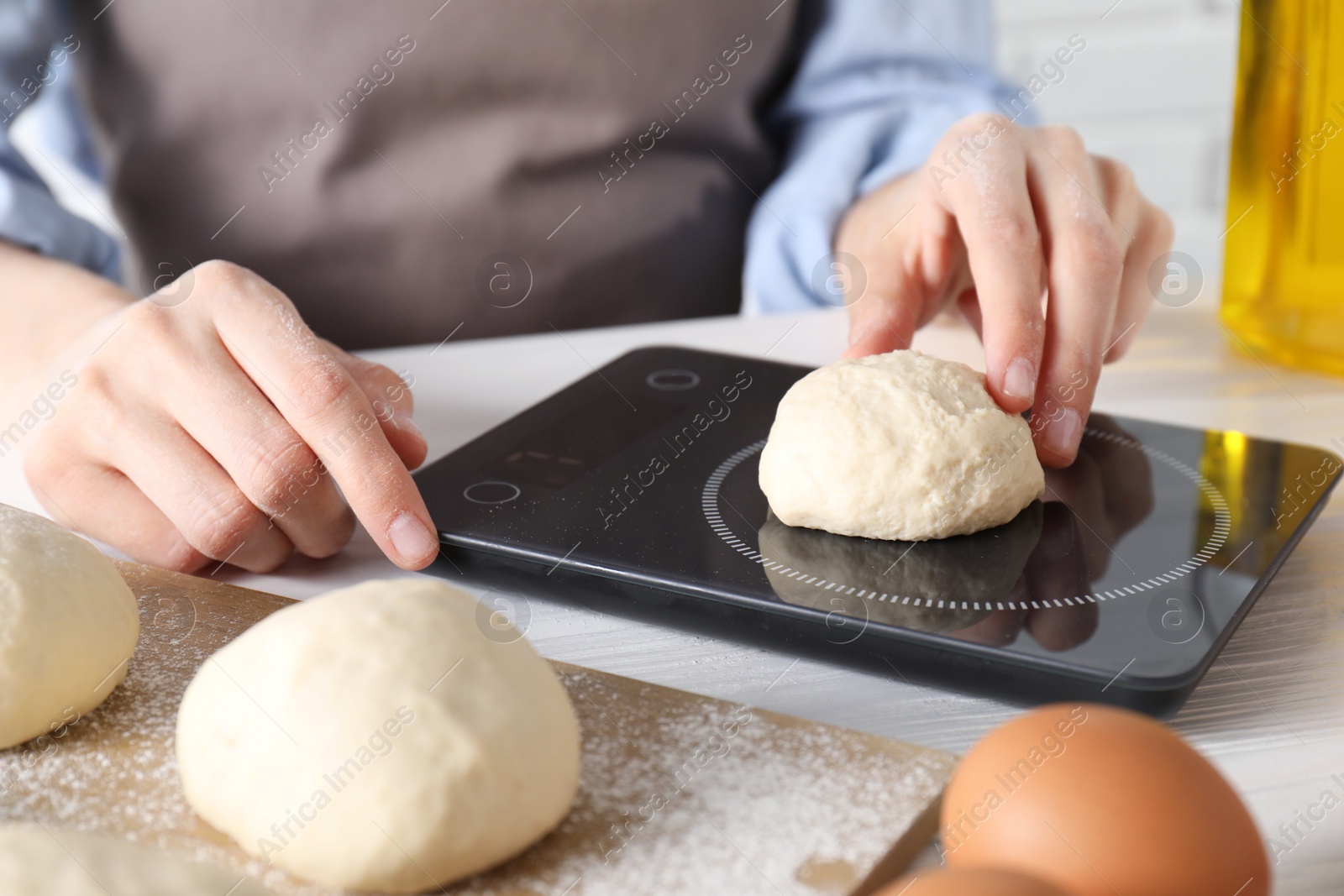 Photo of Woman weighing raw dough ball on kitchen scale at white wooden table, closeup