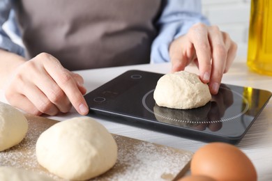 Woman weighing raw dough ball on kitchen scale at white wooden table, closeup