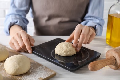 Photo of Woman weighing raw dough ball on kitchen scale at white wooden table, closeup