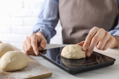 Woman weighing raw dough ball on kitchen scale at white wooden table, closeup