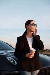 Photo of Smiling woman in sunglasses with paper cup of drink near car outdoors