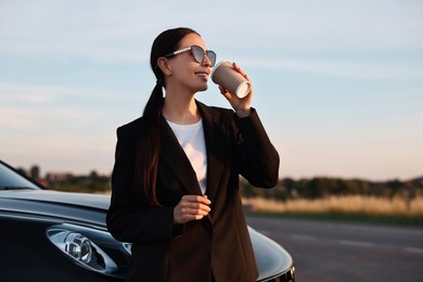 Woman in sunglasses drinking from paper cup near car outdoors