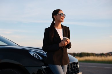 Smiling woman in sunglasses with paper cup of drink near car outdoors