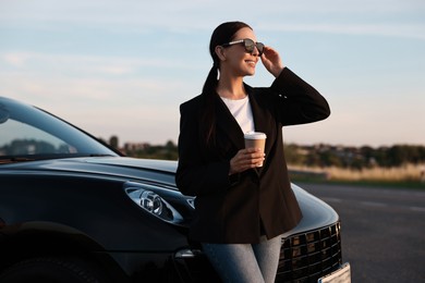 Photo of Smiling woman in sunglasses with paper cup of drink near car outdoors