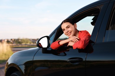 Smiling young woman leaning out of car window, view from outside. Enjoying trip