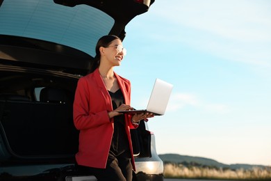 Smiling young woman using laptop near car outdoors, space for text