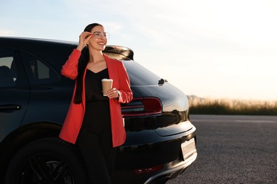 Smiling young woman with paper cup of drink near car outdoors