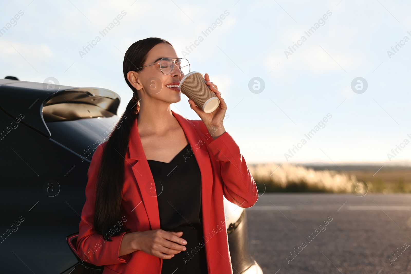 Photo of Beautiful young woman drinking from paper cup near car outdoors, space for text