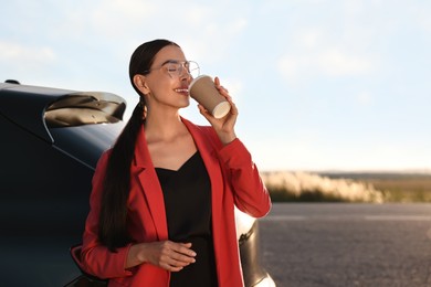 Beautiful young woman drinking from paper cup near car outdoors, space for text