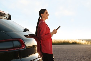 Smiling young woman with smartphone near car outdoors, space for text