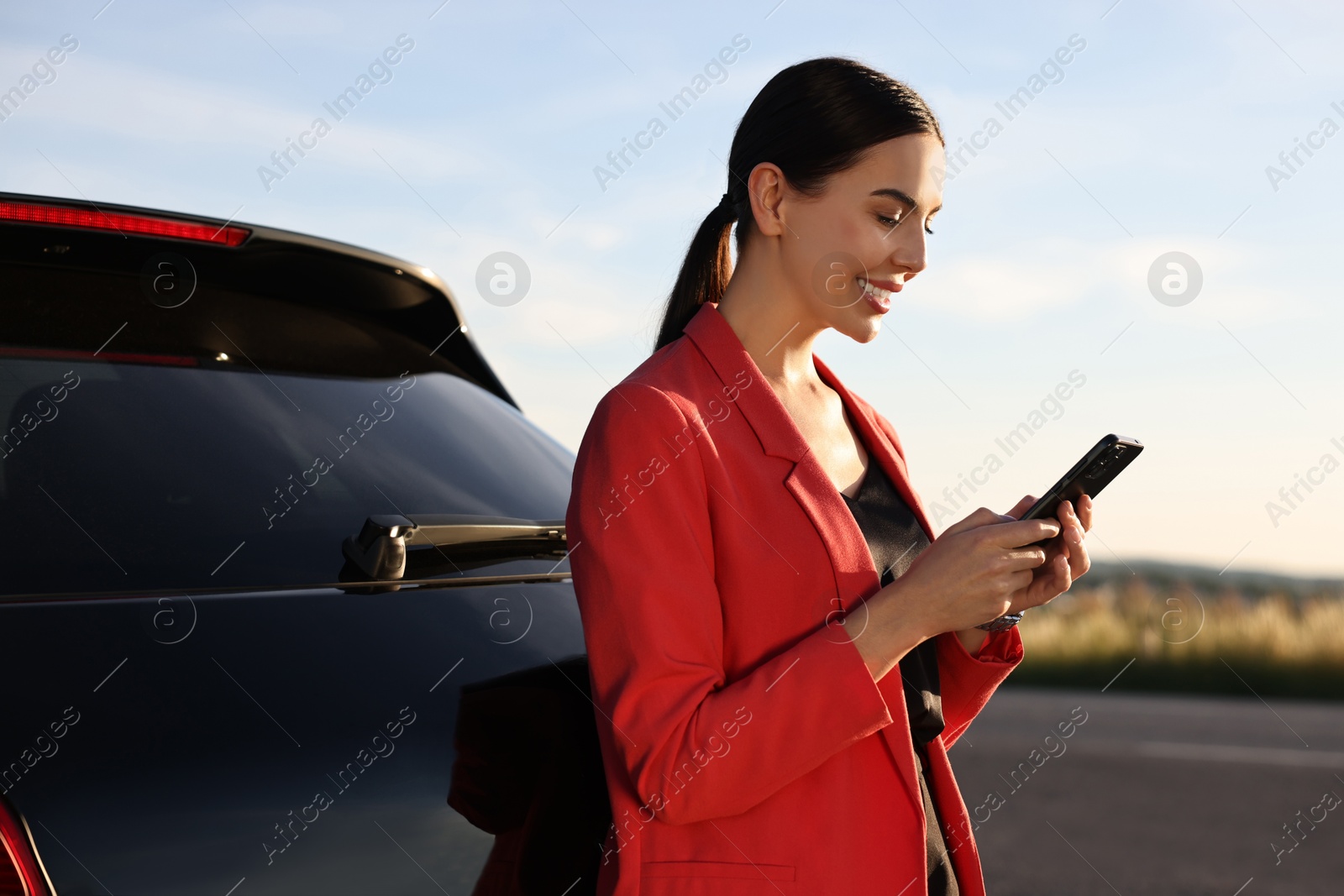 Photo of Smiling young woman with smartphone near car outdoors