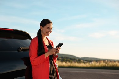 Photo of Smiling young woman with smartphone near car outdoors, space for text