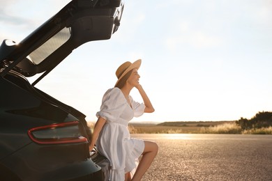 Photo of Smiling young woman in hat near trunk of car at sunset, space for text