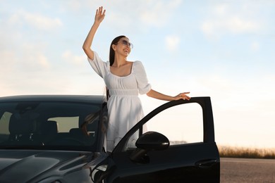 Photo of Smiling young woman in sunglasses leaning out of car outdoors. Enjoying trip