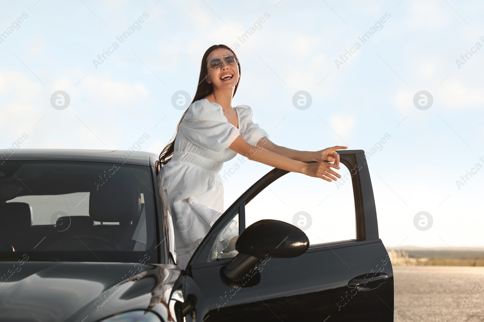 Photo of Smiling young woman in sunglasses leaning out of car outdoors. Enjoying trip