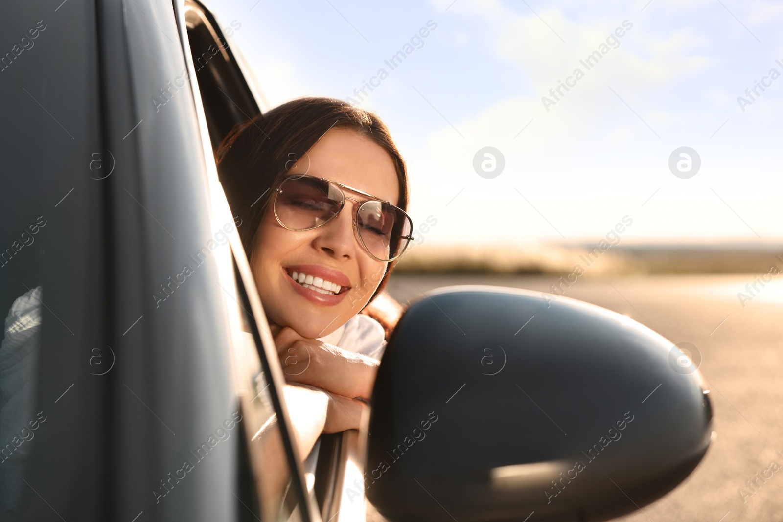 Photo of Smiling young woman in sunglasses leaning out of car window outdoors. Enjoying trip