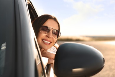 Photo of Smiling young woman in sunglasses leaning out of car window outdoors. Enjoying trip