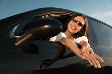 Smiling young woman in sunglasses leaning out of car window outdoors, low angle view. Enjoying trip