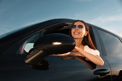 Smiling young woman in sunglasses leaning out of car window outdoors, low angle view. Enjoying trip