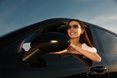 Smiling young woman in sunglasses leaning out of car window outdoors, low angle view. Enjoying trip