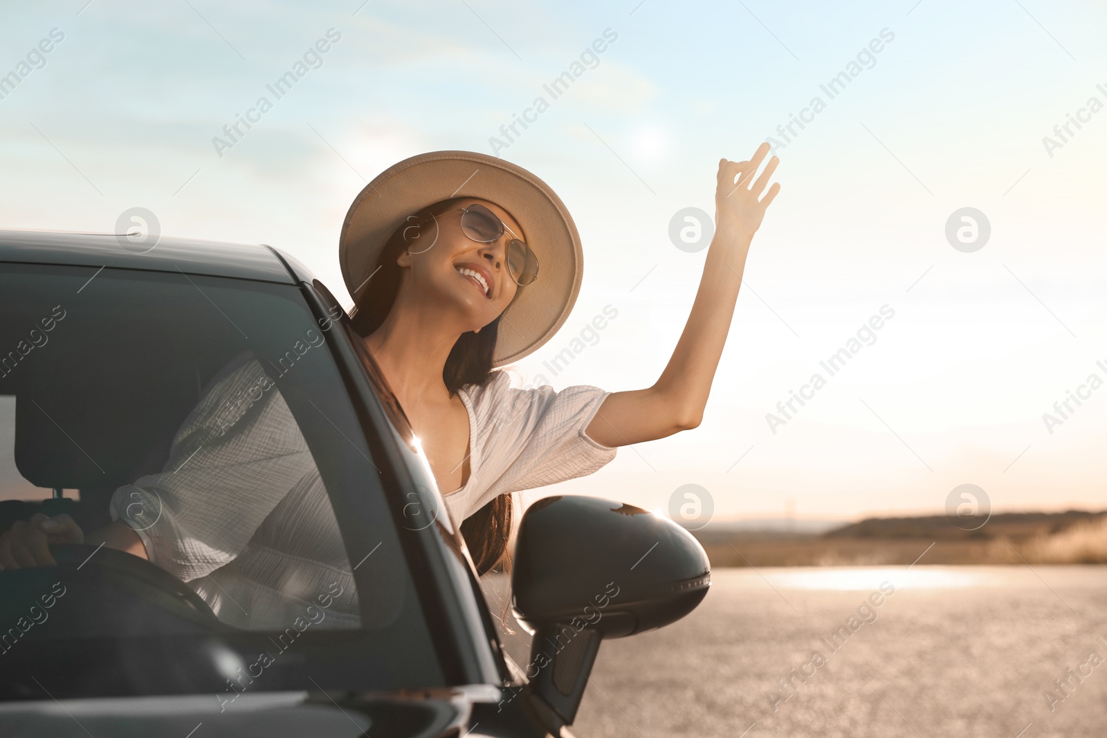 Photo of Smiling young woman in sunglasses with hat leaning out of car window outdoors. Enjoying trip