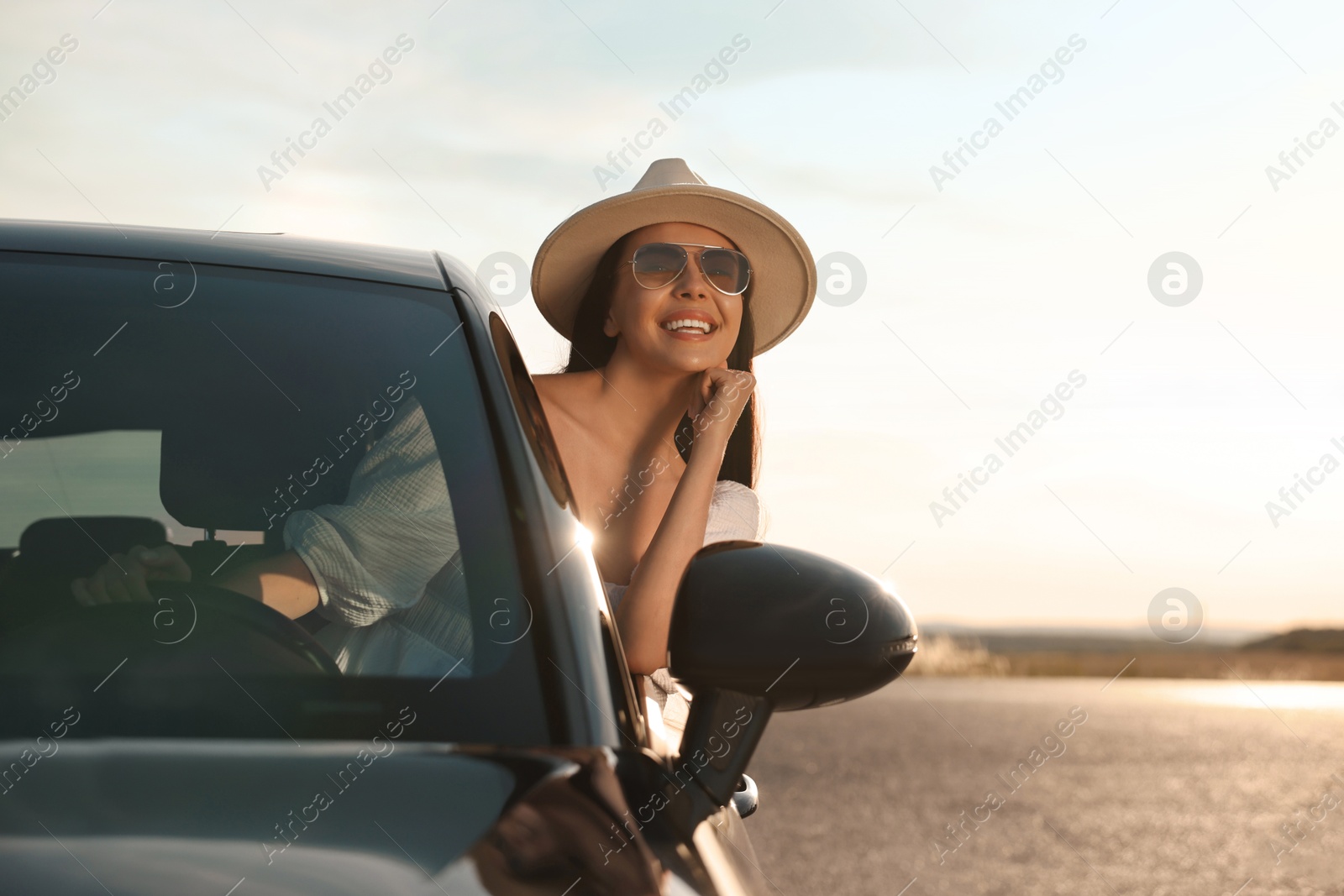 Photo of Smiling young woman in sunglasses with hat leaning out of car window outdoors. Enjoying trip