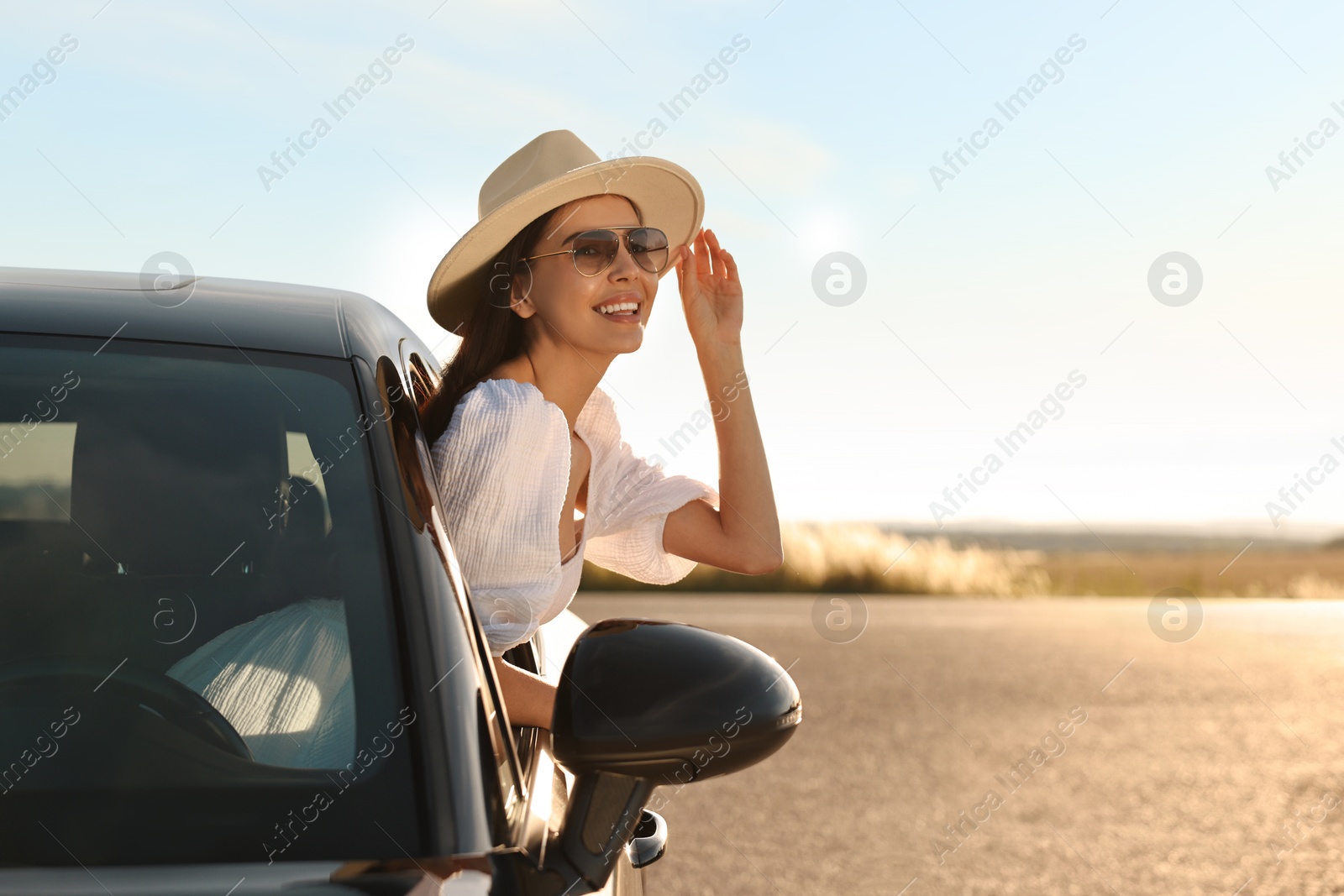 Photo of Smiling young woman in sunglasses with hat leaning out of car window outdoors. Enjoying trip