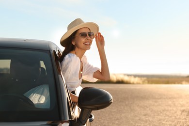 Smiling young woman in sunglasses with hat leaning out of car window outdoors. Enjoying trip