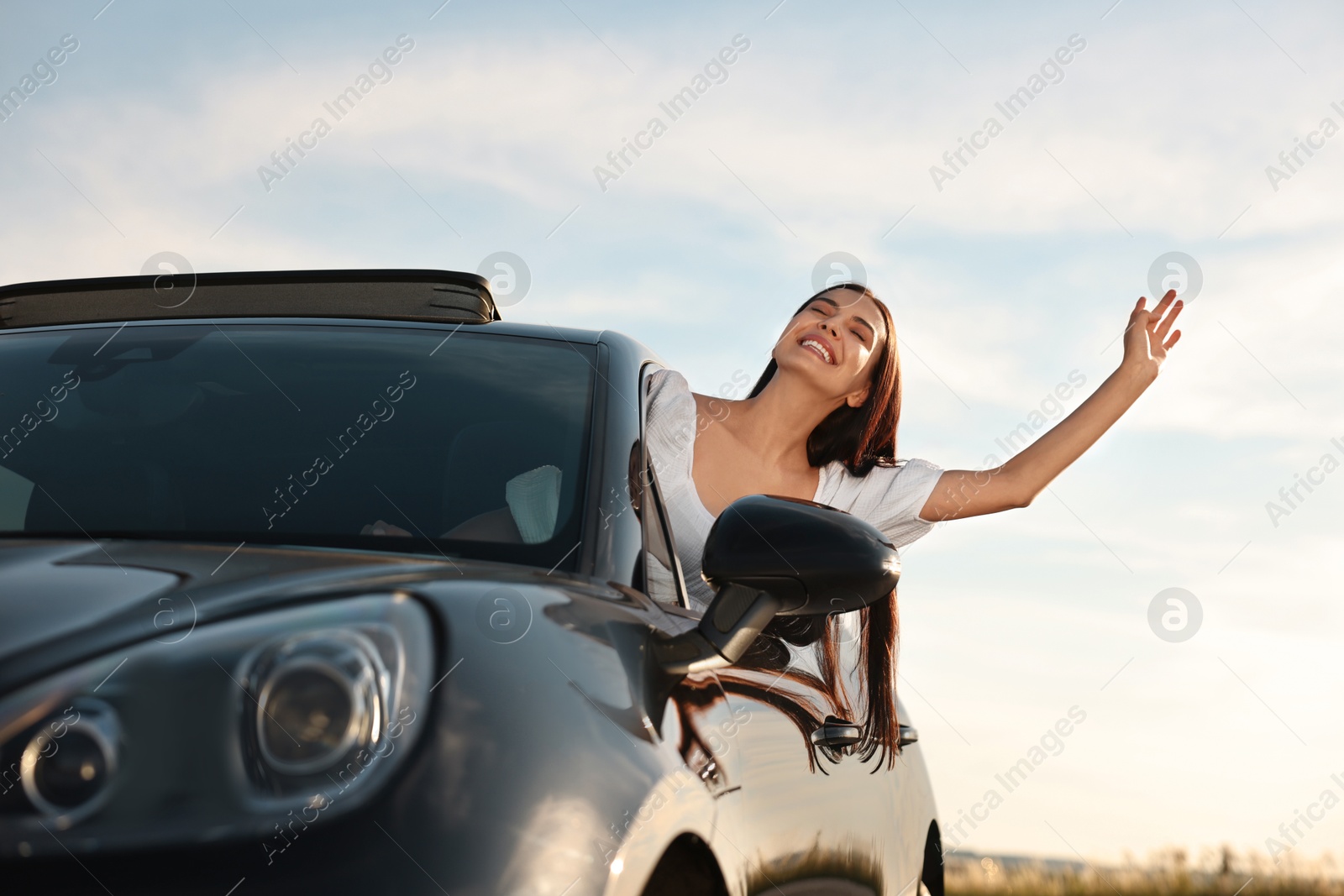 Photo of Smiling young woman leaning out of car window outdoors, low angle view. Enjoying trip