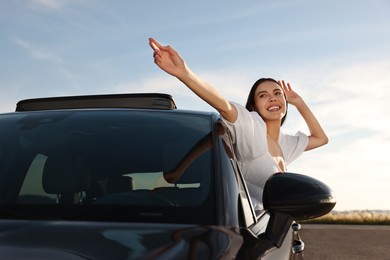 Photo of Smiling young woman leaning out of car window outdoors. Enjoying trip