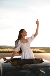 Enjoying trip. Smiling woman leaning out of car roof outdoors