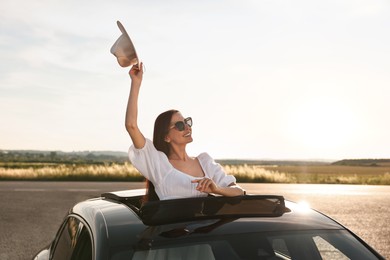 Enjoying trip. Smiling woman in sunglasses with hat leaning out of car roof outdoors