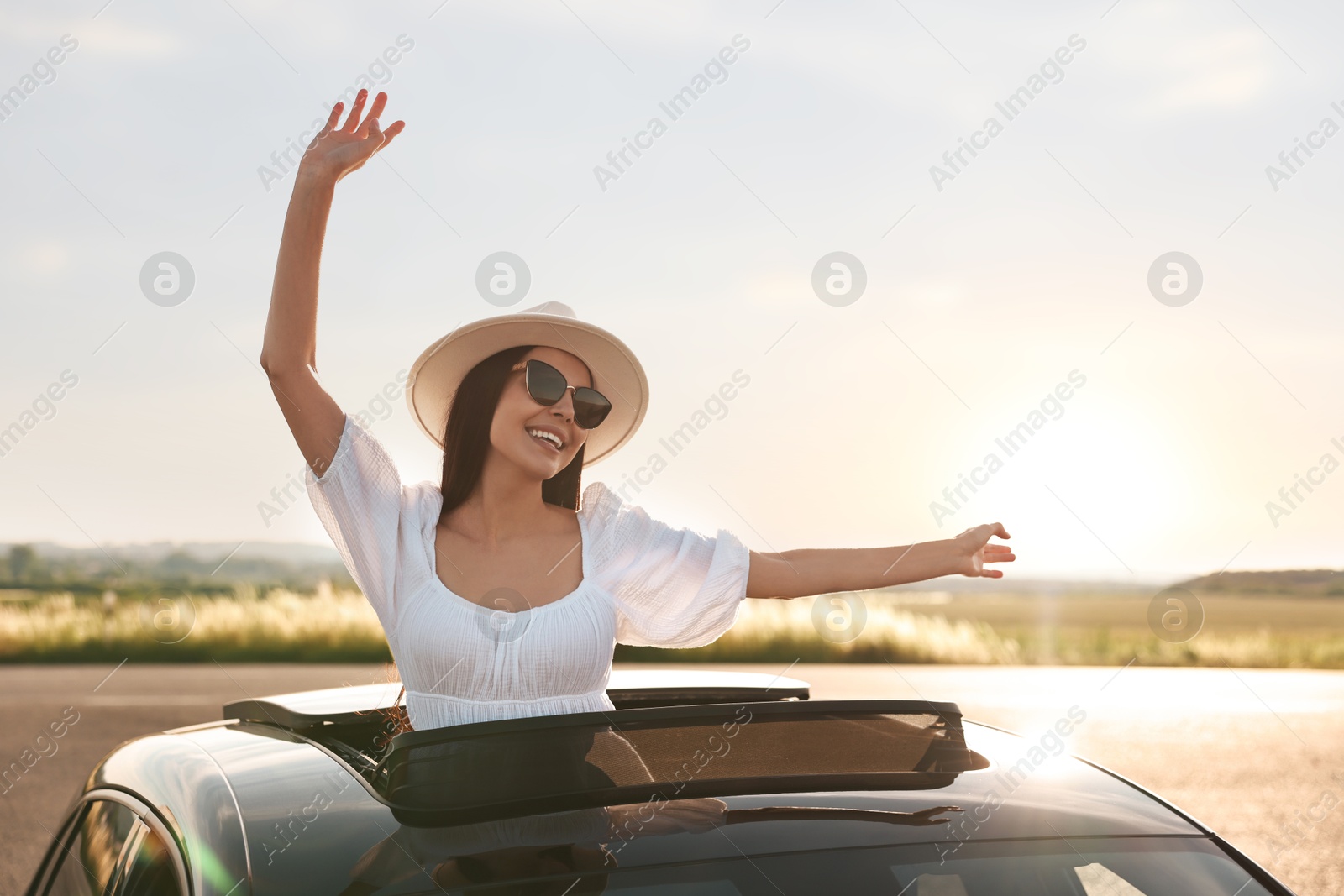 Photo of Enjoying trip. Smiling woman in sunglasses with hat leaning out of car roof outdoors