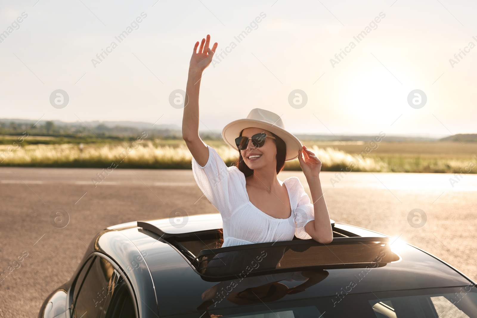 Photo of Enjoying trip. Smiling woman in sunglasses with hat leaning out of car roof outdoors