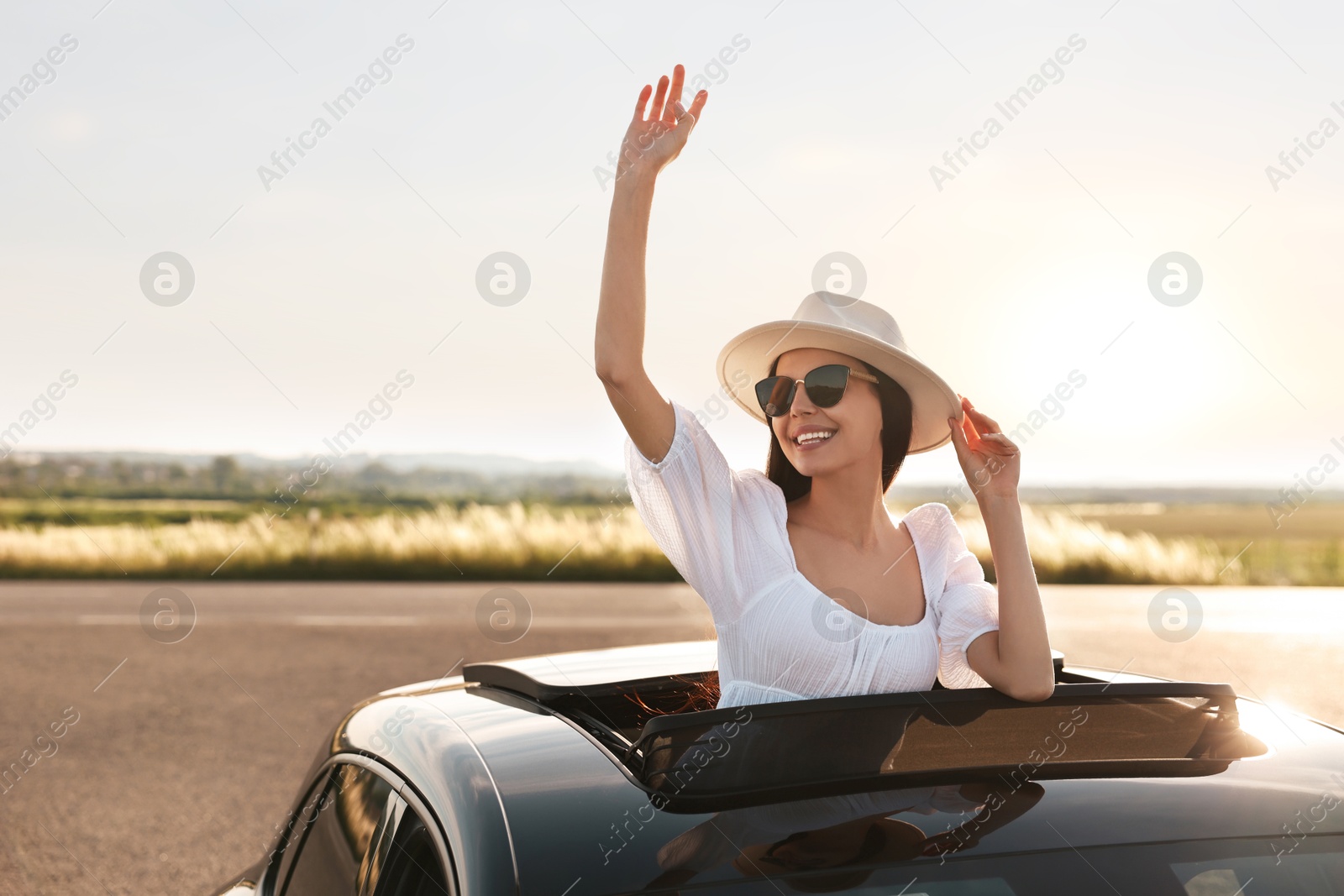 Photo of Enjoying trip. Smiling woman in sunglasses with hat leaning out of car roof outdoors