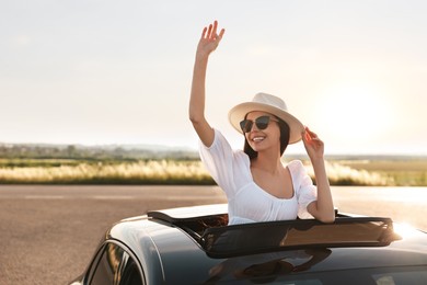 Enjoying trip. Smiling woman in sunglasses with hat leaning out of car roof outdoors