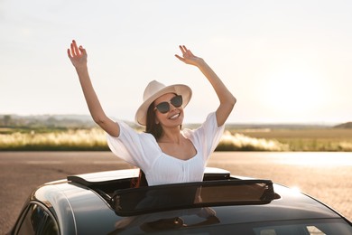 Photo of Enjoying trip. Smiling woman in sunglasses with hat leaning out of car roof outdoors