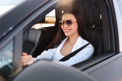 Smiling young woman in sunglasses with seatbelt driving car, view from outside