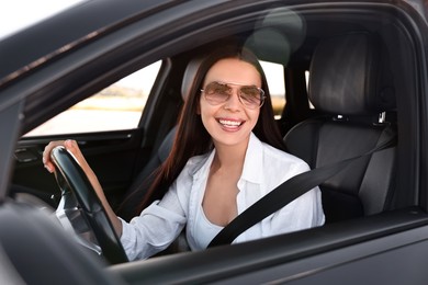 Photo of Smiling young woman in sunglasses with seatbelt driving car, view from outside