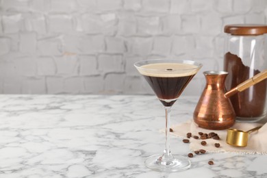 Refreshing cocktail in glass, beans, cezve and ground coffee on white marble table, space for text