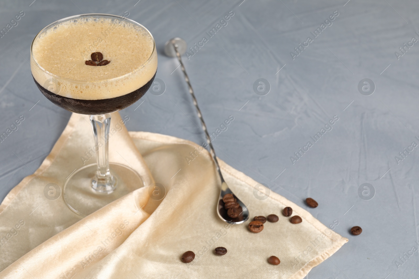 Photo of Fresh coffee cocktail in glass, beans and mixing spoon on grey table. Space for text