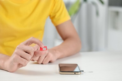 Man holding SIM card near smartphone at white wooden table indoors, closeup. Space for text