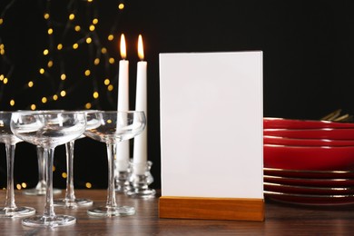 Photo of Menu holder, burning candles and clean dishware on wooden table against blurred lights