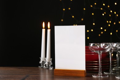 Photo of Menu holder, burning candles and clean dishware on wooden table against blurred lights, space for text