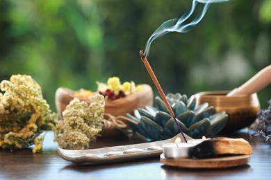 Photo of Incense stick smoldering in holder, candles and dry flowers on wooden table outdoors