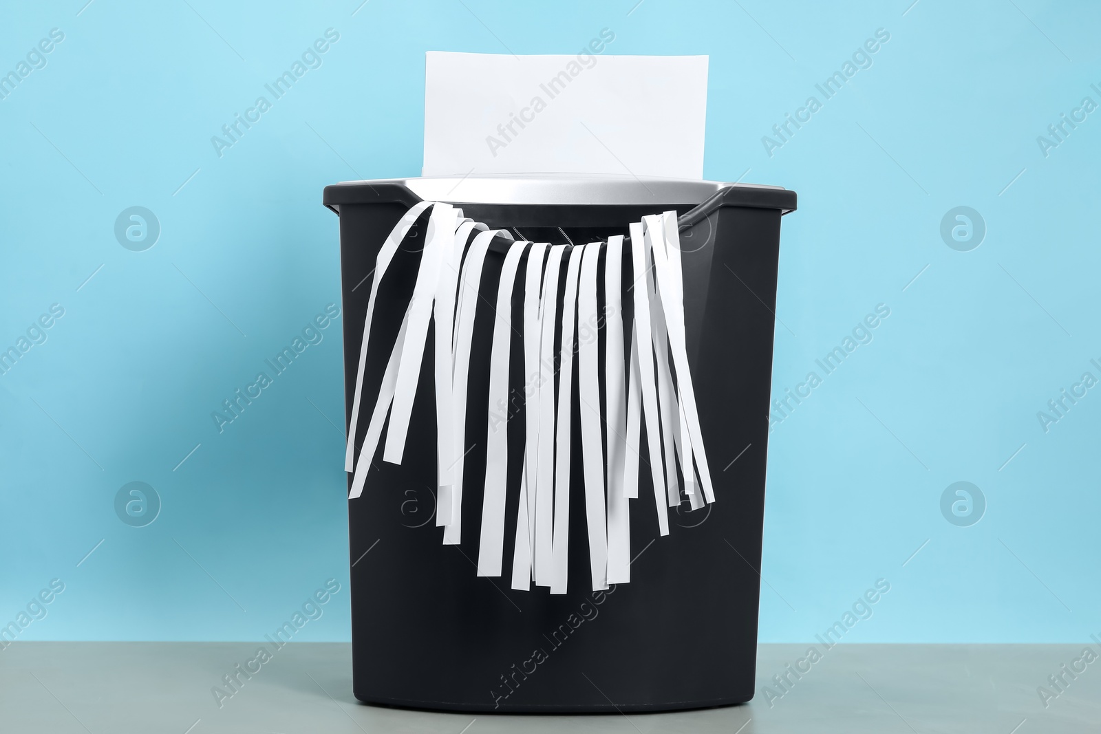 Photo of Destroying sheet of paper with shredder at grey table against light blue background