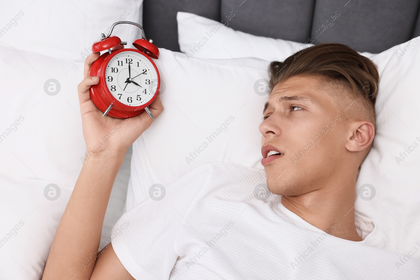 Photo of Sleepy young man in bed with alarm clock waking up at lunch time